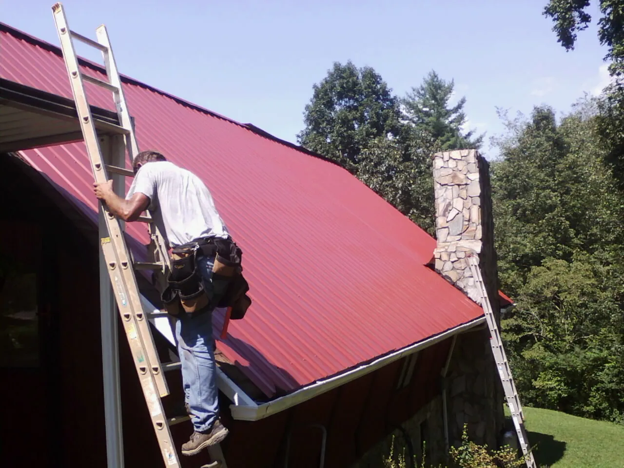 Roofer on a ladder beside a red metal roof and stone chimney — Soffit & Fascia Repair by Roofing Brothers in Gilmer